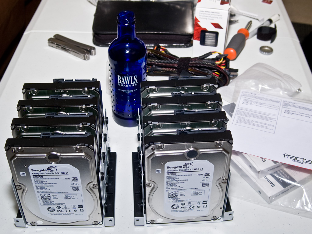 A table with eight hard drives in the foreground and some manuals to the side, a bottle of Bawls energy drink, some cabling, and miscellaneous tools and components in the back.